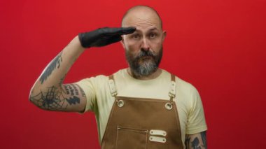 Kahverengi tulumlu kel bir adam canlı kırmızı bir arka plana karşı eldivenli bir elle selam veriyor. Kendine güveni ve tarzını yansıtıyor..