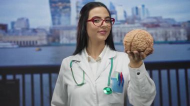 Kadın doktor, deniz kenarında steteskopla gezerken, güvenle gülümseyen bir beyin modeli tutuyor..