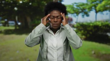 Parkta kafası tutulan bir kadın arka planda yeşillik olan bir ortamda stresli görünüyor..