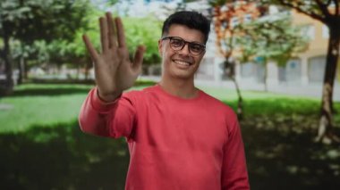 Açık hava parkında gülümseyen gözlüklü genç adam güneşli bir günde kırmızı kazak giyiyor. Etrafı yemyeşil ve şehir planlarıyla çevrili. Keyifli bir ortam izlenimi veriyor.