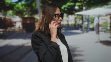 Güneşli bir şehir sokak ortamında telefonla konuşan, gözlüklü ve siyah ceketli, modern şehir yaşam tarzını ve iletişimini İspanyol bağlamında sergileyen bir kadın..