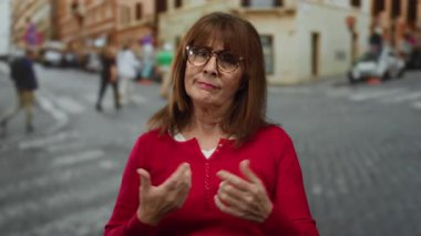 Kırmızı kazaklı yaşlı İspanyol kadın hareketleriyle kalabalık bir şehir caddesinde canlı şehir yaşamını resmediyor..