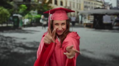 Pembe mezuniyet elbisesi içindeki genç kadın, hareketli bir şehir caddesinde canlı bir şehir ortamında başarı ve başarı sergileyen parmağıyla kutlama yapıyor..