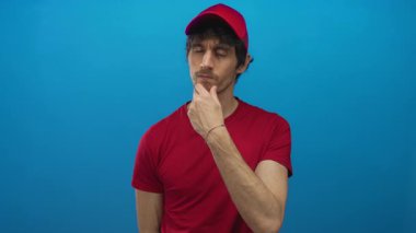 Kırmızı şapkalı ve gömlekli genç teslimatçı, mavi stüdyoda parmağını yukarı kaldırıyor. Düşünceli bir ifadeyle..