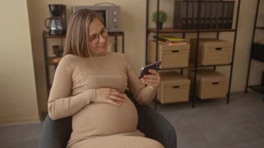 Ofiste hamile bir kadın akıllı telefona bakarken gülümsüyor. Arkasında dosyalar ve kutular var. Modern bir iş ortamı öneriyor..