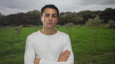 Kolları çapraz duran genç adam, ağaçlar ve bulutlu gökyüzü ile yeşil parkta duruyor, beyaz bir gömlek ve küpeler giyiyor, açık havada düşünceli bir ifade yayıyor..