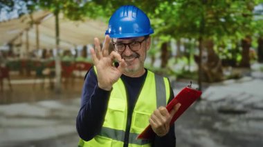 Miğfer ve güvenlik yeleği giyen bir adam park ortamında, şehir ortamında, açık hava panosuyla tamam işareti yapıyor. İnşaat bağlamında olgun bir güven gösteriyor..