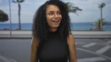 Sahilde yürürken gülümseyen gözlüklü bir kadın. Arka planda palmiye ağaçları ve okyanus var. Mavi bir gökyüzünün altında..