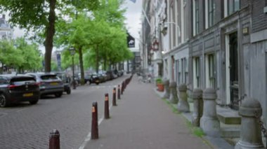 Park etmiş arabaların, kaldırımların, ağaçlarla çevrili caddelerin ve yeşil yapraklı bir gölgelik altında geleneksel mimarinin olduğu Amsterdam caddesinin odaksız görüntüsü..