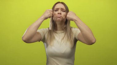 Sarışın genç bir kadın, izole edilmiş sarı arka plan duvarına karşı komik bir ağlama hareketi yapıyor. Yüzüne yakın yumrukları kapalı, neşeli ve abartılı bir ifade sergiliyor..