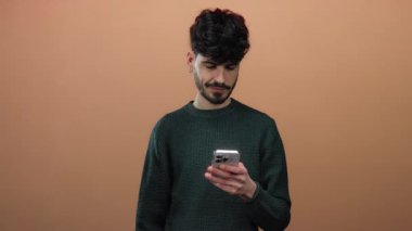 Telefonlu genç adam kahverengi arka planda kameraya bakıp gülümsüyor, izleyicilerle bağlantı kuruyor, cazibesini ve teknolojisini gösteriyor..