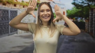 Mavi gökyüzünün altındaki şehir sokağında elleri çerçeveli gülümseyen kadın sıradan bir şehir ortamında mutluluk ve güven sergiliyor..