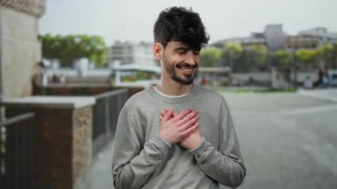 Sokak ortamında göğsünde elleri göğsünde sıcacık gülümseyen genç bir adam..
