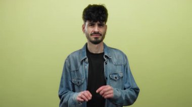 Tereddüt ve belirsizliği vurgulayan, parlak sarı bir arkaplan önünde kot ceket giyen, şüpheci bir ifadeyle isteksizliğini ifade eden genç bir adam..