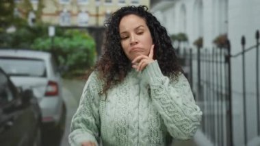 Yeşil kazaklı, kıvırcık saçlı genç bir kadın, arka planda eski binalar ve park halindeki arabalarla şehir caddesinde düşünceli bir şekilde kafa yoruyor.
