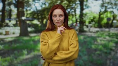 Düşünceli bir şekilde açık hava parkında duran, kırmızı saçlı sarı bir kazak giyen, yemyeşil ve dingin doğayla çevrili bir kadın..