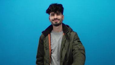 İzole edilmiş mavi arka planda rahat ceketli genç adam olumlu ve başarılı olduğunu ima ederek gülümsüyor.