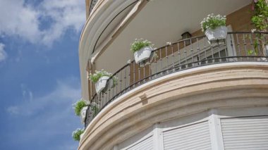 Güneşli bir günde İspanya 'da saksı bitkileriyle dolu Akdeniz cephesinde, berrak mavi gökyüzünün altında çiçeklerle süslenmiş eğri büğrü bir bina balkonu yer alıyor..