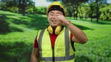 Güneşli bir günde parkta gülümseyen güvenlik yeleği ve kasketli Asyalı bir adam..