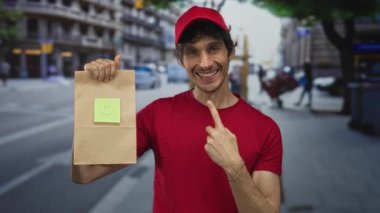 Kırmızı şapkalı ve kırmızı tişörtlü teslimatçı, işlek şehir caddesinde gülen yüzlü kağıt torba ve işaret parmağı tutuyor..