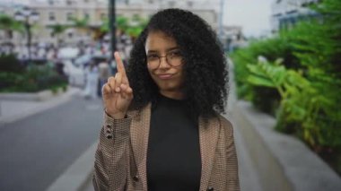 Genç İspanyol kadın şehir caddesinde kameraya işaret parmağıyla işaret ediyor ve arkasında bulanık bir kalabalık var..