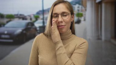 Sarışın genç bir kadın kenar mahallede yanağa dokunuyor, dedikodu gizliliği fısıldıyor..