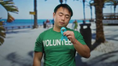 Mavi kurdeleli ve gönüllü tişörtlü genç adam güneşli bir sahil yürüyüşünde duruyor, sakin su ve yeşillikle deniz kenarında kanser farkındalığını vurguluyor..