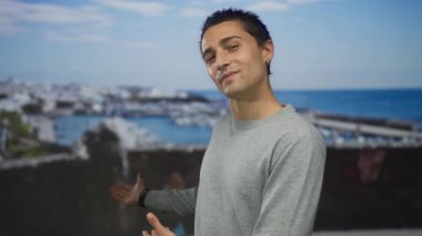 Genç Hispanik adam deniz kenarı liman manzarasını sunmak için kollarını geriye doğru uzatıyor; daveti karşılıyor.