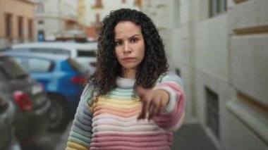 Kıvırcık saçlı, gökkuşağı süveter giyen bir kadın dışarıda kollarını kavuşturup arka planda park etmiş arabalarla şehir caddesinde reddedildiğini gösteriyor..