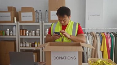 Bağış merkezinde Asyalı bir adam, arka planda malzeme ve giysi raflarıyla çevrili akıllı telefonuyla bağışların etiketlendiği bir kutuda eşyaları kontrol ediyor..