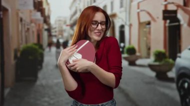 Gözlüklü kadın, şehir caddesinde kalp şeklinde bir hediye kutusu tutuyor. Açık havada verilen özel hediyeye neşe ve minnettarlık gösteriyor..