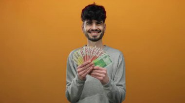 Norveç kronunu izole edilmiş turuncu arka planda tutan adam gülümsüyor, zenginlik ve mutluluk sergiliyor..