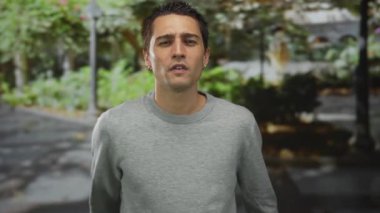 Gri kazak giyen genç İspanyol adam, alacakaranlıkta ağaçlarla kaplı caddede kameraya doğru çıplak orta parmaklarını sallıyor..