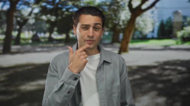 Genç İspanyol bir adam güneşli bir parkta dikiliyor. Yeşil ağaçlar ve gölgeler arasında düşünceli olduğunu gösteriyor.