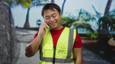 Gündüz vakti şehir merkezinde akıllı telefondan konuşan genç bir adam güvenlik yeleği giyiyor..