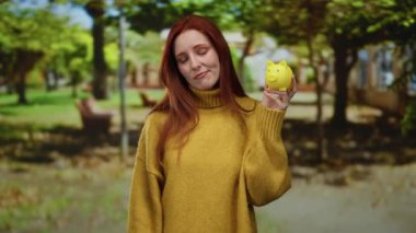 Serene parkında sarı kumbarayı elinde tutan, rahat bir kazak giyen, yeşil ağaçların altında düşünceli ve güneş ışığıyla çevrili bir kadın..