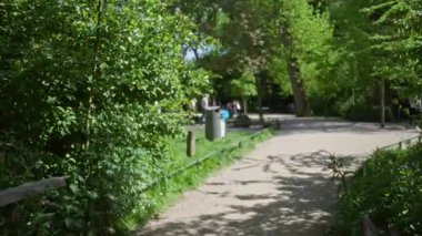 Yeşil ağaçlar ve renkli çiçeklerle çevrili güneşli bir parkta bulanık insanlar, arka planda bokeh etkisi olan sakin bir atmosfer sağlıyor..