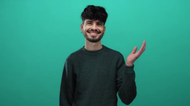 Genç bir adam yeşil bir arka planda gülümseyerek avucunun içini işaret ediyor, kendine güveni ve cazibesi ile süveter giymiş sıradan bir stile sahip..