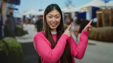 Pembe kazaklı bir kadın kalabalık bir pazar yerini işaret ediyor, insanlarla çevrili ve açık hava tezgahları, canlı alışveriş deneyimi öneriyor..