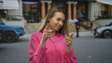 Kadın gülümsüyor ve kentin pozitif enerjisini gösteren parlak pembe bir tişört giymiş bir şekilde kentin caddesini işaret ediyor..