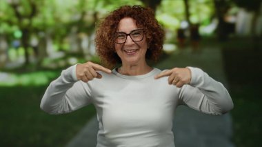 Güneşli bir parkta gülümseyen ve el kol hareketi yapan bir kadın, açık havada, gözlük takıyor ve günlük beyaz kıyafetler giyiyor, bulanık yeşillik ve ağaçlarla çevrili, neşeli bir anı yakalıyor..