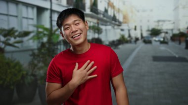 Şehrin sokaklarında neşeli bir şekilde gülen genç bir adam kırmızı tişört giyip açık havada mutluluk ve enerjiyi somutlaştırıyor.