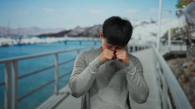 Deniz kenarındaki gezinti alanında gülümseyen ve gözlerini ovuşturan, günlük giysiler giyen, manzaralı bir plaj ve arka planda açık mavi gökyüzü olan genç bir adam..