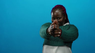 Kadın neşeli bir şekilde poz veriyor. Parlak mavi bir arkaplan karşısında, örgülü saçı ve yeşil kazağıyla kendine güvenini ve eğlencesini ifade ediyor..