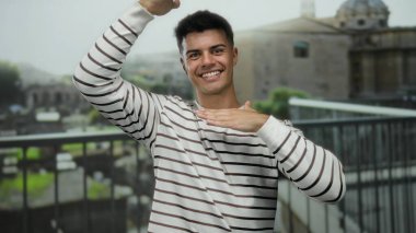 Roma harabelerinin önünde poz veren genç bir adam, gün boyunca açık havada gülümseyen, çizgili bir kazak giyen, kentsel bir ortamda enerjik ve çekici bir enerji sergileyen..