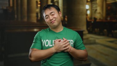 Gönüllü olarak etiketlenmiş yeşil gömlekli genç adam kilisede mutlu duruyor, ellerini göğsüne koyuyor, dini bir ortamda minnettarlık ve memnuniyeti ima ediyor..