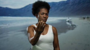 Sahilde el kol hareketleri yapan, arka planda dağlar olan bir kadın açık havaya davet ediyor..