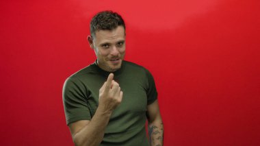 Genç İspanyol bir adam, izole edilmiş kırmızı bir arkaplan karşısında parmağını hareket ettiriyor, kolunda yeşil gömlekli, görünür bir dövmesi olan davetkar bir yüz ifadesi sergiliyor..