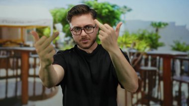 Sakallı genç adam şehir sokağının yanındaki terasta kaba bir jest yapıyor. Arka planda restoran ve kahve var..