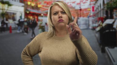 Bej renkli süveter giyen genç sarışın kadın, şehir caddesine karşı elini kalçasında gezdirerek parmak işareti yapıyor..